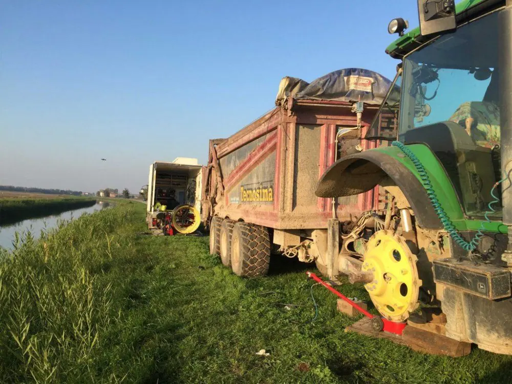 Un trattore con rimorchio fermo su una strada di campagna, con la ruota del rimorchio smontata
